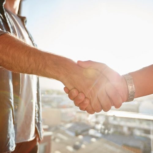 Closeup of two shaking male hands with sun flare. Focus on handshake against cityscape.